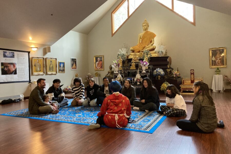 a group of students sits on the floor of a Buddhist temple with a "nunk" wearing a red cloak