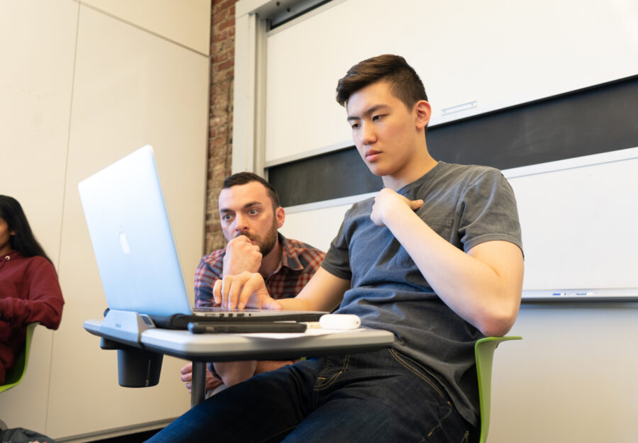 male student sitting at a desk typing on a laptop with male instructor looking over his shoulder