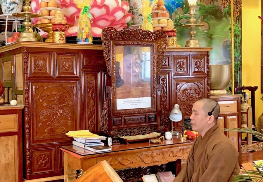 A Buddhist monk in a saffron robe sits on the floor playing a drum in front of a temple display