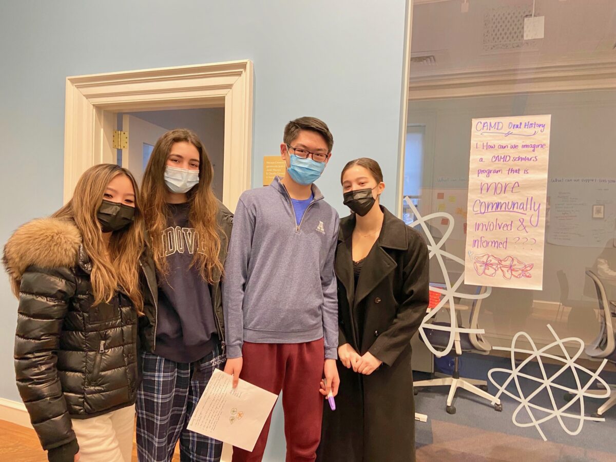 four Workshop students looking at the camera standing next to a list they made on the wall