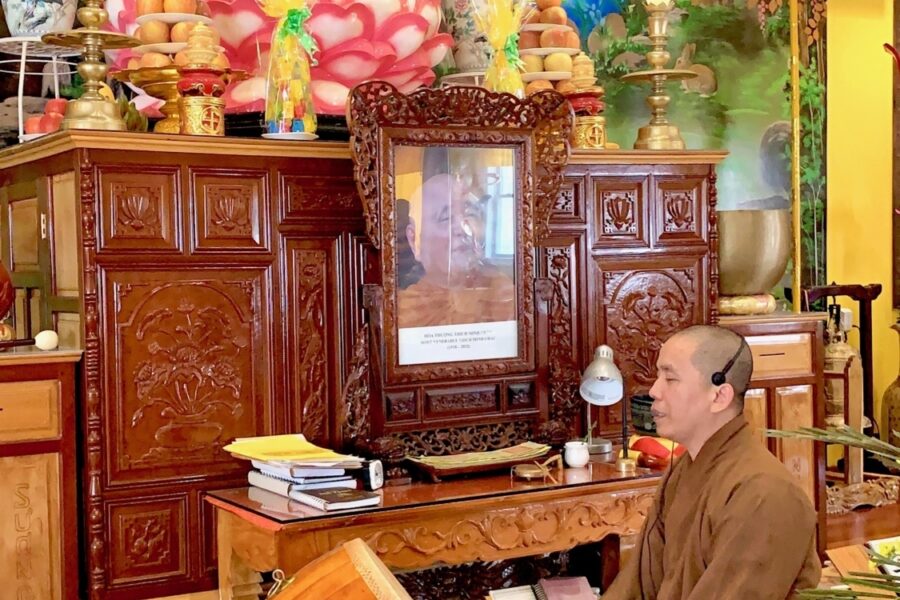 A Buddhist monk in a saffron robe sits on the floor playing a drum in front of a temple display
