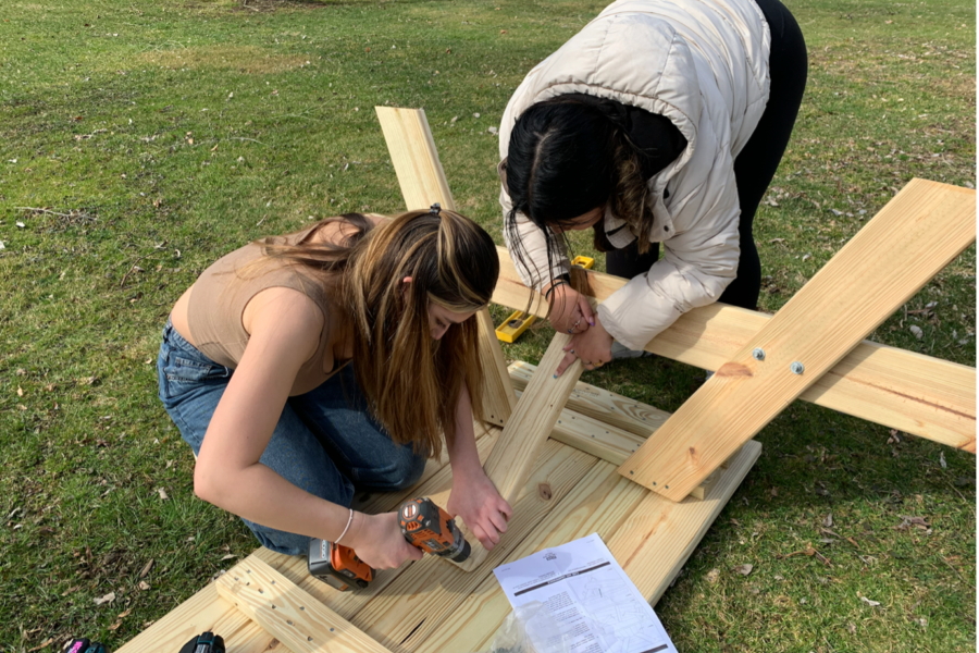 two young teens work outside in winter coats using a drill to put together a picnic table