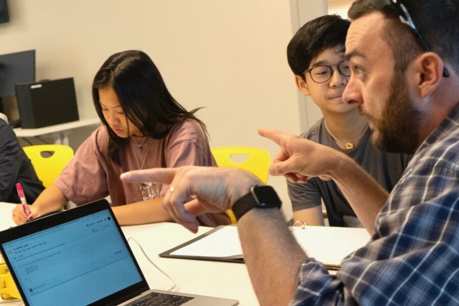 male teacher sitting at a table discussing ethics in a CS classroom with students