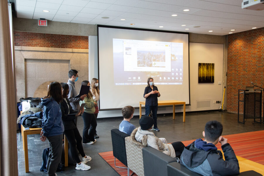 students gathered in large room with an orange rug and a white board; teacher talking in the front of the room
