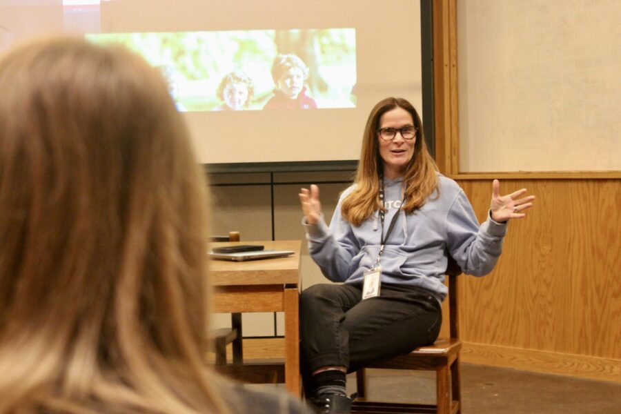 woman sits in a chair in a classroom with both arms spread wide; students sit facing her